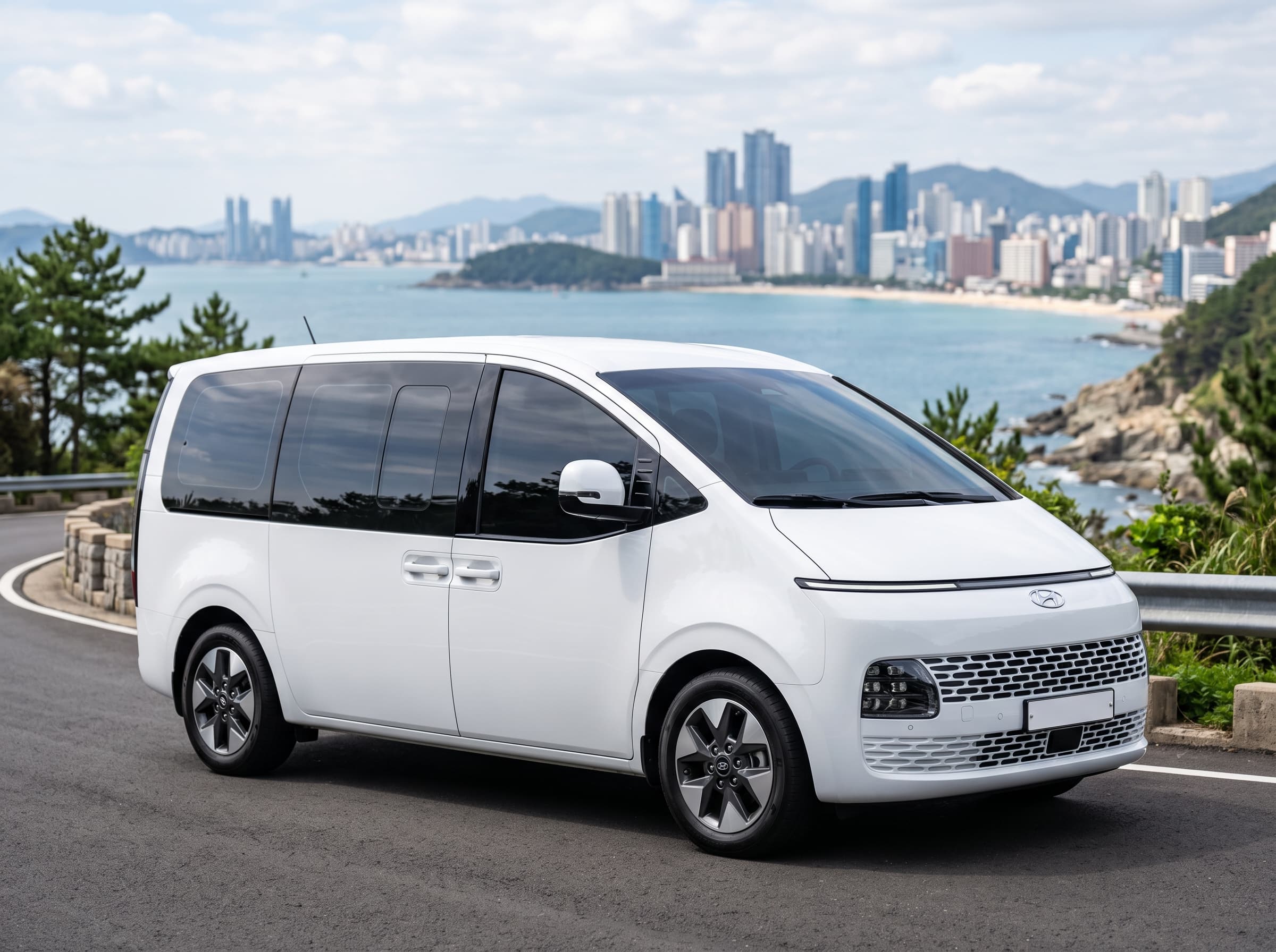 White Hyundai Staria passenger van on a coastal road with the Busan skyline behind it